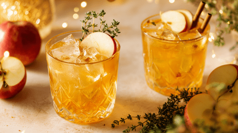 Zwei Gläser mit Apfel-Zimt-Cocktail mit Gin. Darum liegen frische Äpfel und ein Thymianzweig als Dekoration.