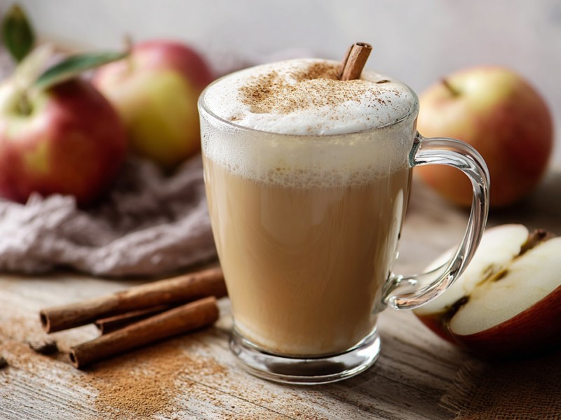 Ein Apple Cider Chai Latte, der auf einem Tisch steht. Daneben sind Zimtstangen, ein aufgeschnittener Apfel und ganz Äpfel zu sehen.