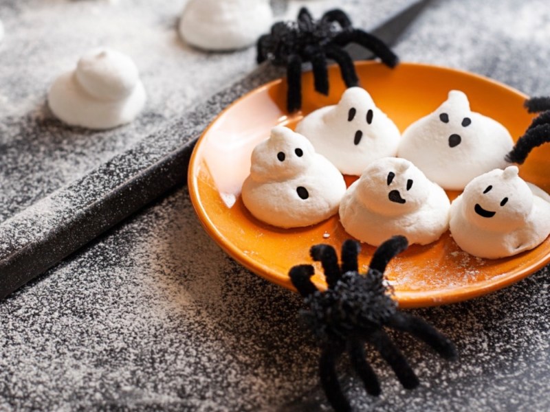 Fünf süße Baiser-Geister auf einem orangen Teller. Spinnen als Halloween-Deko liegen daneben.