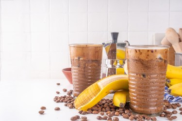 Zwei Gläser mit Bananen-Kaffee-Smoothie. Eine Banane und Kaffeebohnen liegen als Dekoration außen herum.