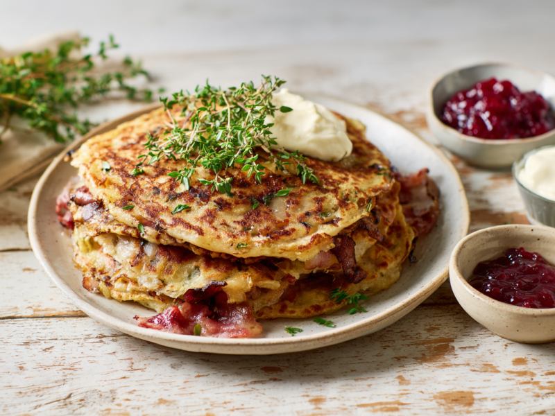 Mehrere Birnen-Speck-Pfannkuchen auf einem Teller. Sie sind mit Thymian und Crème fraîche serviert. Neben dem Teller stehen Schälchen mit Preiselbeermarmelade und mehr Crème fraîche.