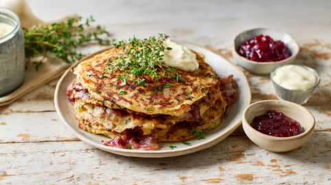 Mehrere Birnen-Speck-Pfannkuchen auf einem Teller. Sie sind mit Thymian und Crème fraîche serviert. Neben dem Teller stehen Schälchen mit Preiselbeermarmelade und mehr Crème fraîche.