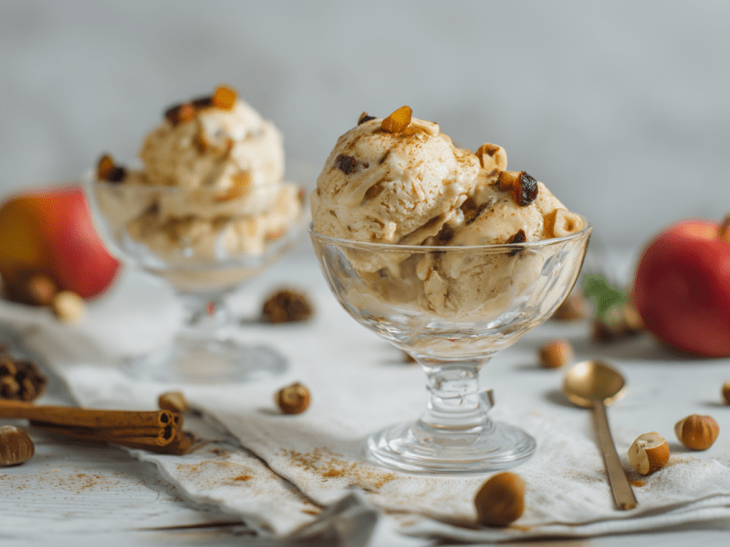 Zwei Schalen Bratapfel-Eis auf einem dekorierten Tisch. Das erste steht im Vordergrund, das zweite weiter im Hintergrund. Auf dem Tisch liegen Äpfel, Zimtstangen und Haselnüsse verteilt.