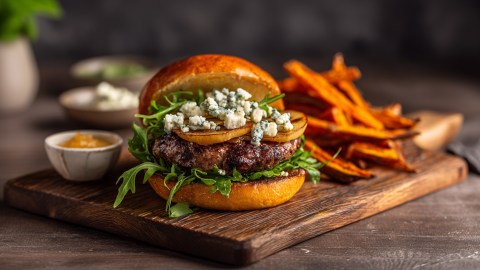 Ein Holzbrett, auf dem ein Blauschimmelkäse mit karamellisierten Äpfeln liegt. Im Hintergrund kann man eine Portion Süßkartoffel-Pommes liegen sehen. Auch Schalen mit Soßen sind zu erkennen.