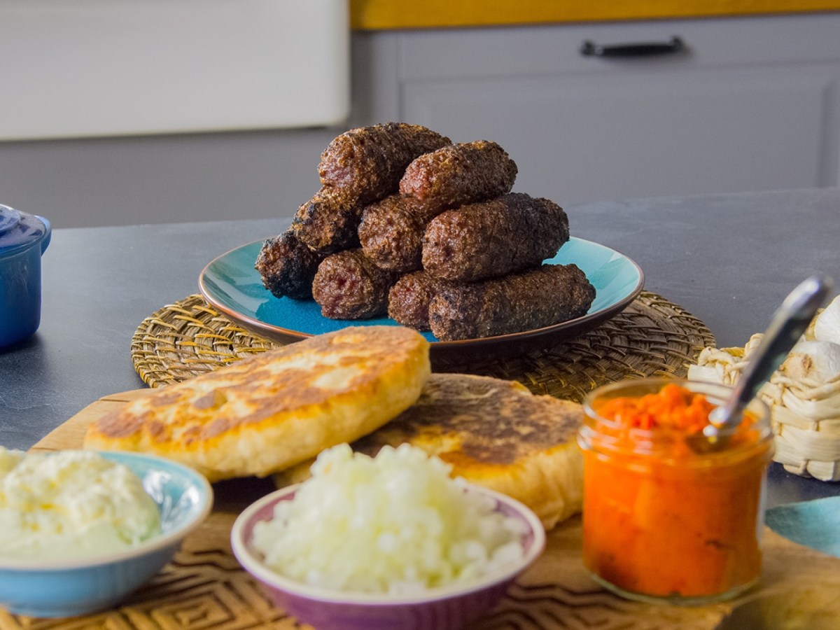 Ein Stapel Cevapcici auf einem Teller im Hintergrund. Vorne sind Fladenbrot und weitere Zutaten zu sehen.