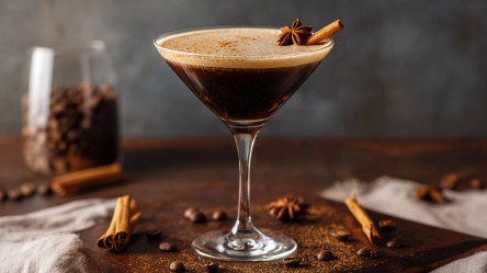 Ein Martini-Glas mit einem Chai Espresso Martini. Das Ganze ist mit einer Zimtstange und Sternanis garniert. Ringsum liegen Zimtstangen und Kaffeebohnen verteilt. Im Hintergrund steht ein Glasgefäß mit Kaffeebohnen.