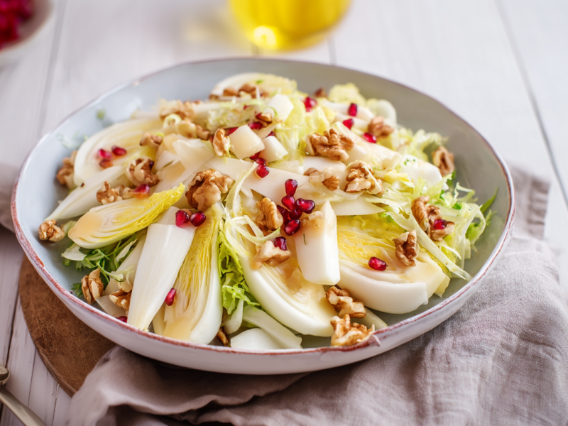 Eine Schale Chicoréesalat mit Apfel auf einem grauen Küchentuch. Links davon liegt eine Gabel. Im Hintergrund links sieht man einige Granatapfelkerne.