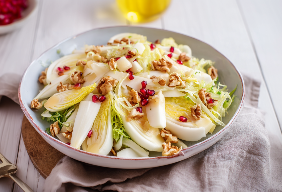 Eine Schale Chicoréesalat mit Apfel auf einem grauen Küchentuch. Links davon liegt eine Gabel. Im Hintergrund links sieht man einige Granatapfelkerne.