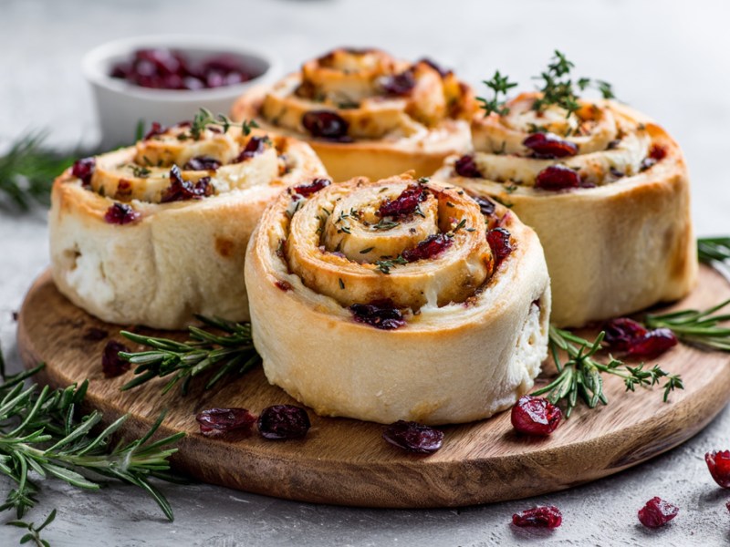 Ein Holzbrett mit Cranberry-Frischkäse-Schnecken. Im Hintergrund steht eine Schale mit getrockneten Cranberrys.