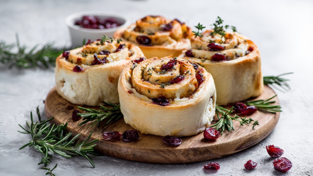 Ein Holzbrett mit Cranberry-Frischkäse-Schnecken. Im Hintergrund steht eine Schale mit getrockneten Cranberrys.