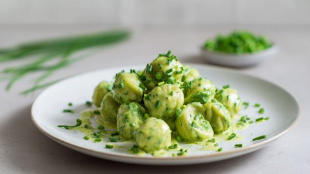 Ein Teller mit einer Portion Erbsen-Pasta mit Frischkäse-Senfsoße. Im Hintergrund kann man einen Bund Schnittlauch und eine Schale mit Erbsen erkennen.