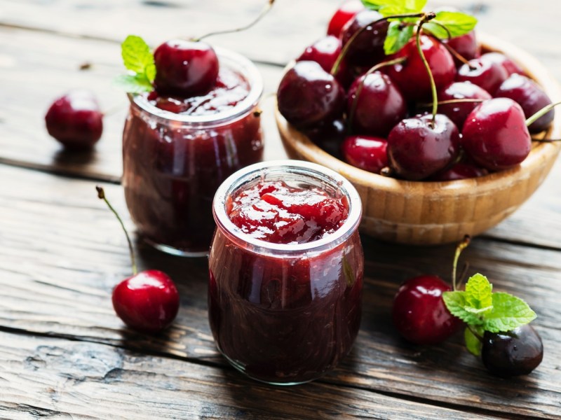 Zwei Gläser mit Espresso-Kirsch-Marmelade. Eine Schale mit frischen Kirschen liegt daneben, ebenso wie einzelne frische Kirschen.