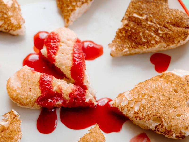 Ein Pancake in Herzform, der in der Mitte zerteilt ist und mit roter Fruchtsoße beträufelt ist. Das Ganze soll ein gebrochenes Herz symbolisieren. Drum herum liegen noch einige weitere kleine Pancakes in Herform.