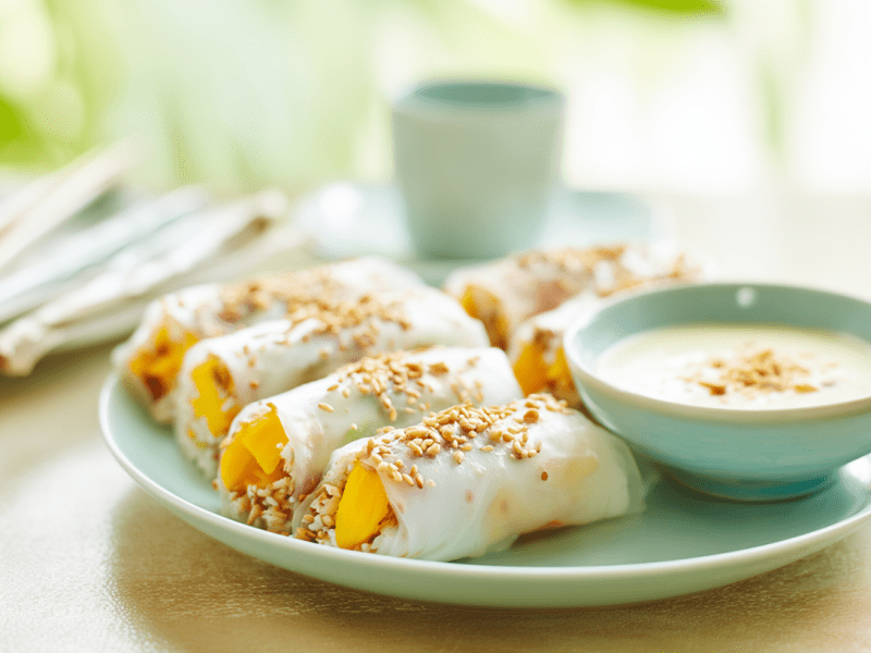Ein Teller mit Frühlingsrollen mit Mango und Klebreis. Daneben steht eine Schale mit Dip.