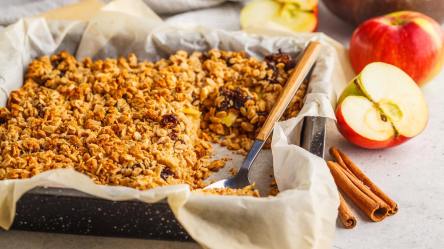 Gebackene Apfelkuchen-Haferflocken in einer Auflaufform, daneben eine Zimtstange und Äpfel.