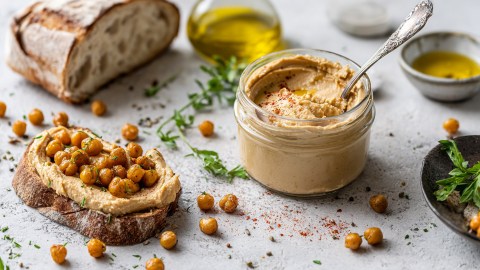 Ein Glas mit gerösteter Kichererbsenbutter, in dem ein Löffel steckt. Daneben liegt eine Scheibe Sauerteigbrot mit gerösteter Kichererbsenbutter und gerösteten Kichererbsen. Im Hintergrund ist ein halber Laib Brot zu sehen. Es kullern Kichererbsen durch die Gegend. Auch ein Gefäß mit Olivenöl, frische Kräuter sind zu sehen.