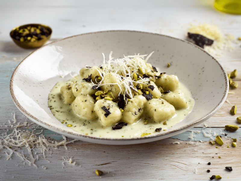Gnocchi mit Pistaziencreme auf einem grauen Teller. Vorne links vor dem Teller liegt frisch geriebener Parmesan. Hinten links ein Schälchen mit Gewürzen. Rechts vom Teller liegen gehackte Pistazien.