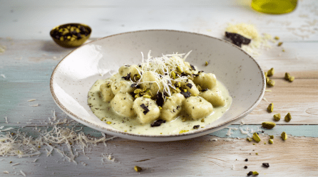 Gnocchi mit Pistaziencreme auf einem grauen Teller. Vorne links vor dem Teller liegt frisch geriebener Parmesan. Hinten links ein Schälchen mit Gewürzen. Rechts vom Teller liegen gehackte Pistazien.
