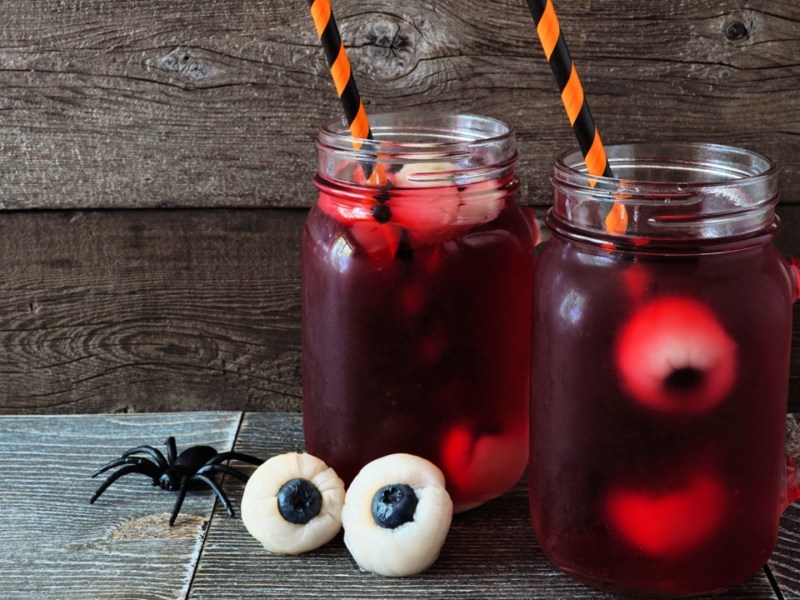 Zwei Gläser mit blutroter Halloween-Bowle ohne Alkohol. Vor den Gläsern liegen zwei Litschi-Heidelbeer-Augen und eine Plastikspinne. Die Getränke sind mit bunten Strohhalmen dekoriert.