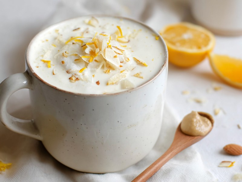 Eine Tasse heiße weiße Schokolade mit Orangen und Mandel, die mit Orangenabrieb und Mandelblättchen garniert ist. Daneben liegt ein Holzlöffel mit Mandelmus. Außerdem ist eine halbe Orange sowie ein Orangenschnitz zu sehen.