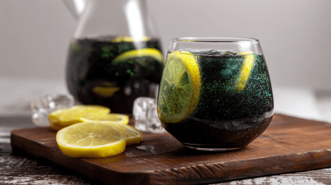 Ein Glas Hexen-Limonade steht auf einem Holzbrett. Daneben liegen einige Zitronenscheiben. Im Hintergrund ist noch eine Karaffe mit der Limo.