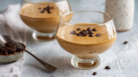 Zwei Dessertgläser mit Hopjesvla, einem holländischen Kaffee-Karamell-Pudding. Beide Puddings sind mit Kaffeebohnen garniert. Links daneben ist eine kleine Schale mit Kaffeebohnen und einem Löffel darin zu sehen.