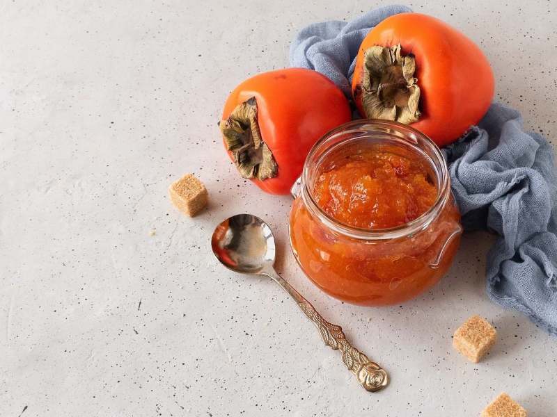 Ein Glas mit Kaki-Marmelade, daneben Zuckerwürfel und ganze Früchte.