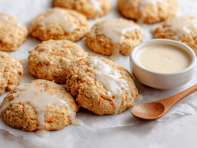Mehrere Karottenkekse liegen auf einem weißen Stück Backpapier. Sie sind mit einer Glasur überzeugen, die in einem Schälchen bei ihnen steht. Daneben liegt ein kleiner Holzlöffel.