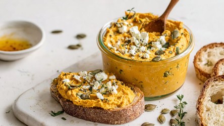 Eine Glasschale mit Kürbis-Feta-Aufstrich. Daneben liegt eine Scheibe, die mit dem Aufstrich bestrichen ist. Ringsum liegen frische Kräuter und geröstete Kürbiskerne herum.