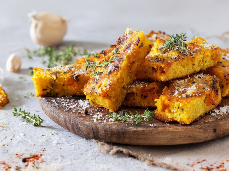Ein rundes Holzbrett mit einigen Kürbis-Polenta-Schnitten, die aufeinander gestapelt sind. Sie sind mit geriebenem Parmesan und frischem Thymian garniert. Ringsum um und im Hintergrund kann man ein paar Kürbisstücke, eine Knoblauchknolle sowie frische Kräuter und Gewürze verstreut liegen sehen.