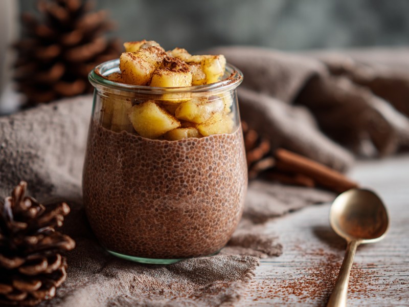 Ein kleines Glas mit Lebkuchen-Chiapudding, der mit Apfelkompott getoppt ist. Ringsum kann man Tannenzapfen, Zimtstangen und ein drapiertes Tuch erkennen.