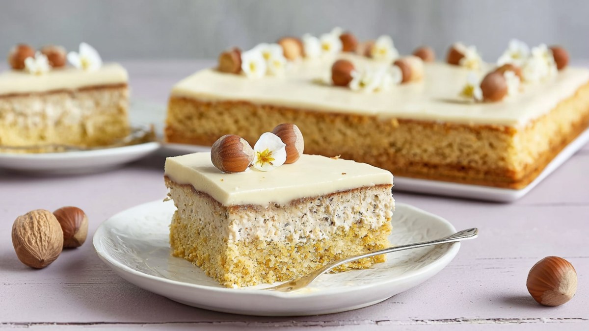 Ein Teller mit einem Stück Lübecker Marzipantorte vom Blech. Es ist mit ganzen Haselnüssen und einer weißen Blüte garniert. Im Hintergrund kann man einen ganzen Blechkuchen sehen.