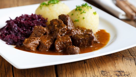 Malzbiergulasch mit Knödeln und Rotkohl auf einem weißen Teller.