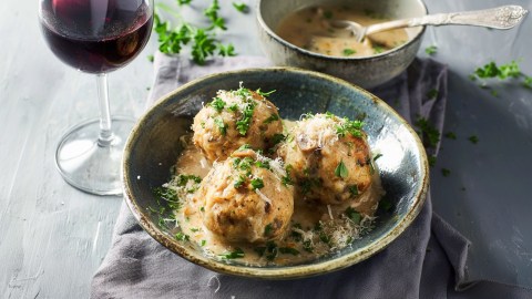 Ein Teller mit drei Maronen-Knödel, die mit Petersilie und Parmesan garniert ist. Dabei steht ein Glas Rotwein und eine Schale mit Rahmsoße.