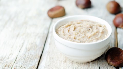 Eine Schale Maronenbutter auf einem hellen Holzuntergrund. Drum herum sind Maronen.