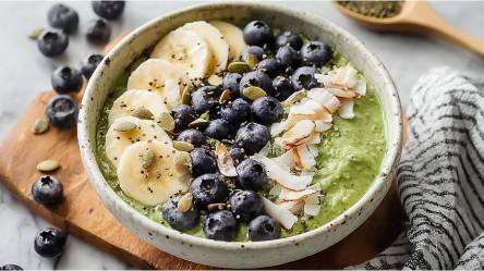 Schale mit Matcha-Overnight-Oats mit Kokos, Bananenscheiben, Kokoschips, Heidelbeeren und Kürbissamen als Topping