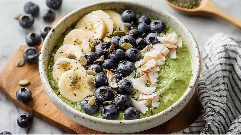 Schale mit Matcha-Overnight-Oats mit Kokos, Bananenscheiben, Kokoschips, Heidelbeeren und Kürbissamen als Topping