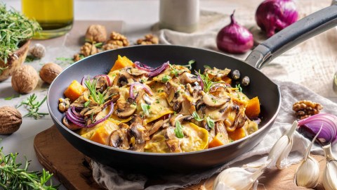 Eine Maultaschen-Pfanne mit Kürbis und Pilzen. Ringsum liegen Knoblauchzehen, Zwiebeln und Walnüsse verteilt.
