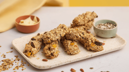 Ein Brett mit Müsli-Käsestangen. Daneben steht ein Schälchen mit Kernen, im Hintergrund ein zweites mit Butter.