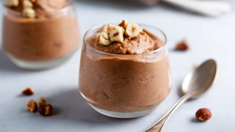 Ein Dessertglas mit Nutella-Mousse. Daneben liegt ein Silberlöffel. Ringsum liegen Haselnüsse herum.