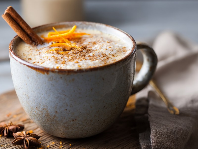 Eine Tasse Orange-Zimt-Latte, die mit einer Zimtstange und Orangenzeste garniert ist. Die Tasse steht auf einem Holzbrett, im Hintergrund ist ein Leinentuch drapiert.