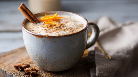 Eine Tasse Orange-Zimt-Latte, die mit einer Zimtstange und Orangenzeste garniert ist. Die Tasse steht auf einem Holzbrett, im Hintergrund ist ein Leinentuch drapiert.