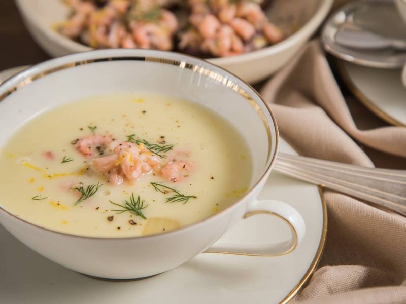 Eine Suppenschale mit ostfriesischer Krabbensuppe, die auf einem Unterteller steht, auf dem auch ein Löffel liegt. Im Hintergrund ist noch eine Schüssel mit Krabben zu sehen.