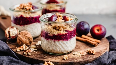 Mehrere kleine Gläser mit Pflaumen-Crumble-Chiapudding, die auf einem Holzbrett platziert sind. Außerdem liegen Zimtstangen, einige Walnüsse und Pflaumen herum.