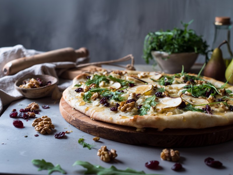 Eine Pizza mit Birne und Cranberrys, die auf einem runden Holzbrett liegt. Ringsum liegen Walnüsse und Cranberrys verstreut. Im Hintergrund sind ein altes Nudelholz, Birnen, eine Schüssel mit Rucola und ein Glasgefäß zu erkennen.