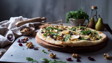 Eine Pizza mit Birne und Cranberrys, die auf einem runden Holzbrett liegt. Ringsum liegen Walnüsse und Cranberrys verstreut. Im Hintergrund sind ein altes Nudelholz, Birnen, eine Schüssel mit Rucola und ein Glasgefäß zu erkennen.