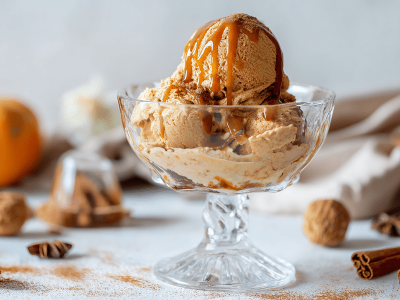 Eine Schale mit Pumpkin-Spice-Eis. Um die Schale verteilt ist Zimt verteilt. Im Hintergrund sieht man einen Kürbis sowie diverse Gewürze.