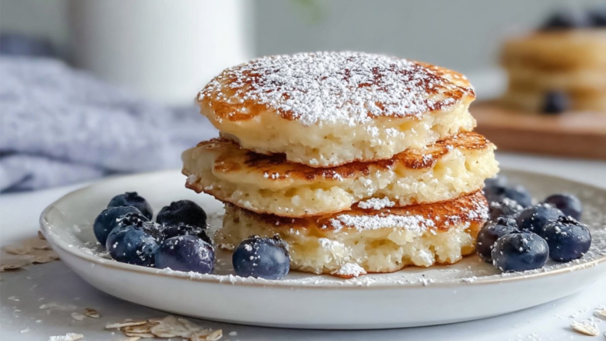 Ein Stapel Quark-Taler mit Haferflocken und Puderzucker auf einem Teller. Drumherum liegen frische Heidelbeeren.