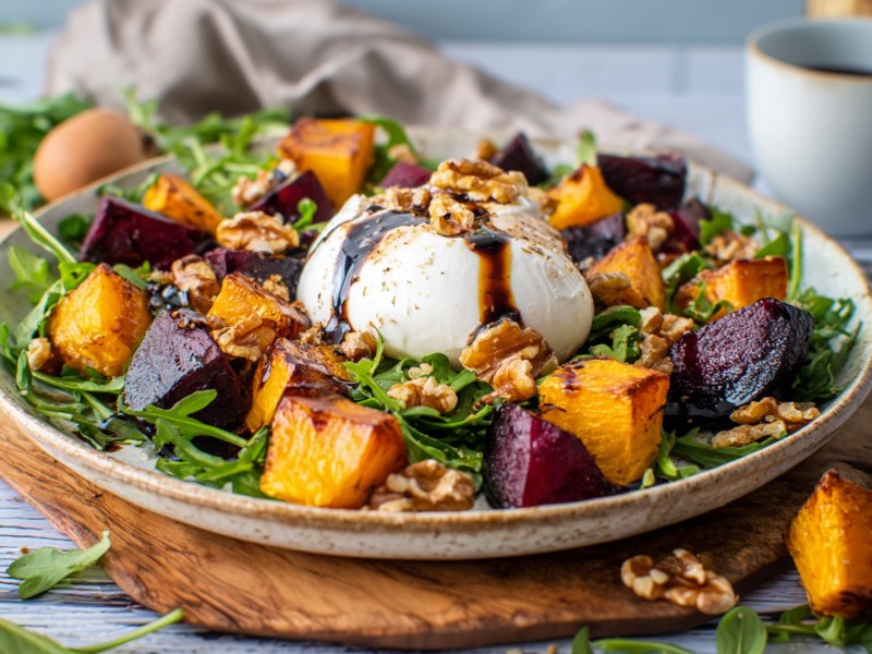 Ein Teller mit Rote-Bete-Kürbis-Salat mit Burrata. Ringsum liegen Kürbiswürfel, Walnüsse und Rucola verstreut. Im Hintergrund steht eine Tasse Kaffee und ein Ei ist zu erkennen.