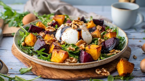 Ein Teller mit Rote-Bete-Kürbis-Salat mit Burrata. Ringsum liegen Kürbiswürfel, Walnüsse und Rucola verstreut. Im Hintergrund steht eine Tasse Kaffee und ein Ei ist zu erkennen.